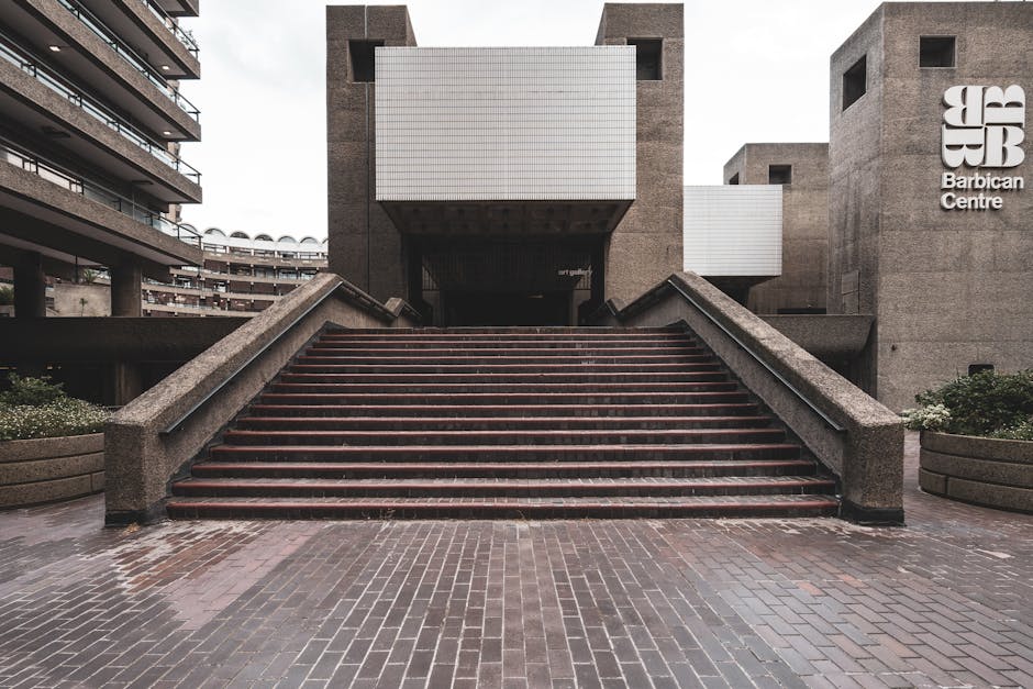 A black and white photograph of the exterior of the Barbican Centre showcasing its modern architectural design with two prominent rectangular brick towers positioned symmetrically in the foreground. T