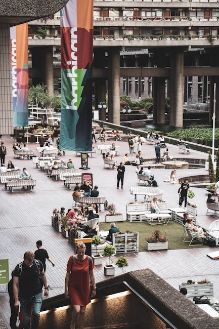 An urban outdoor public space with a paved plaza featuring numerous seating areas, including wooden benches and planters filled with various small shrubs and flowers. Several people are seated, walkin