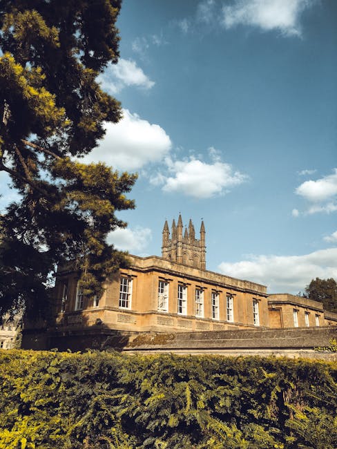 A historic stone building with classical architectural features, including large rectangular windows with white frames, situated in a landscaped area. The structure has a warm, ochre-colored sandstone