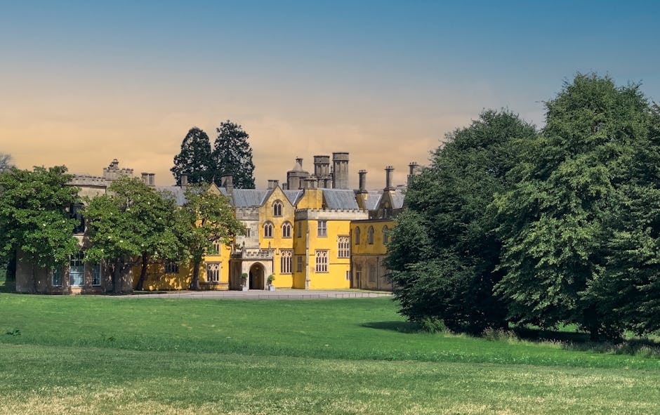 A historic stone building with classical architectural features, including large rectangular windows with white frames, situated in a landscaped area. The structure has a warm, ochre-colored sandstone